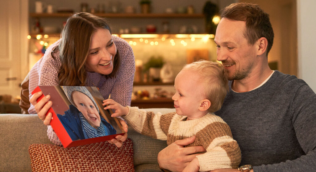 Een gezin zit in een gezellige woonkamer en bekijkt een fotoboek, een kind wijst lachend naar het boek.
