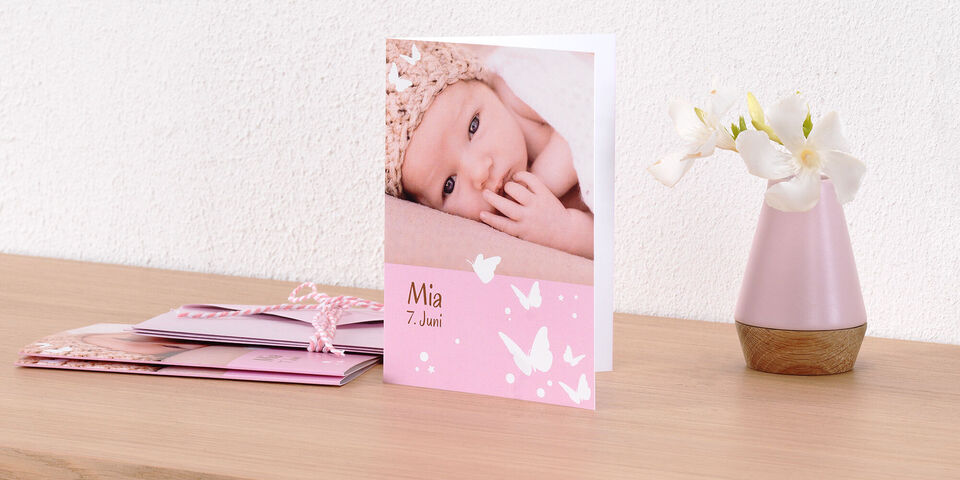 Een delicate, roze wenskaart met een foto van een slapende baby. "Mia 7 juni" staat in liefdevol schrift op de kaart. Naast de kaart staat een decoratieve vaas met bloemen.