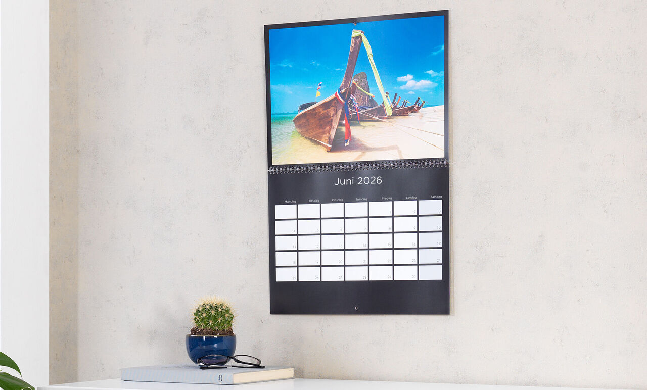 Een wandkalender toont een boot op het strand, met juni 2025 aangegeven. De kalender hangt aan een grijze muur.