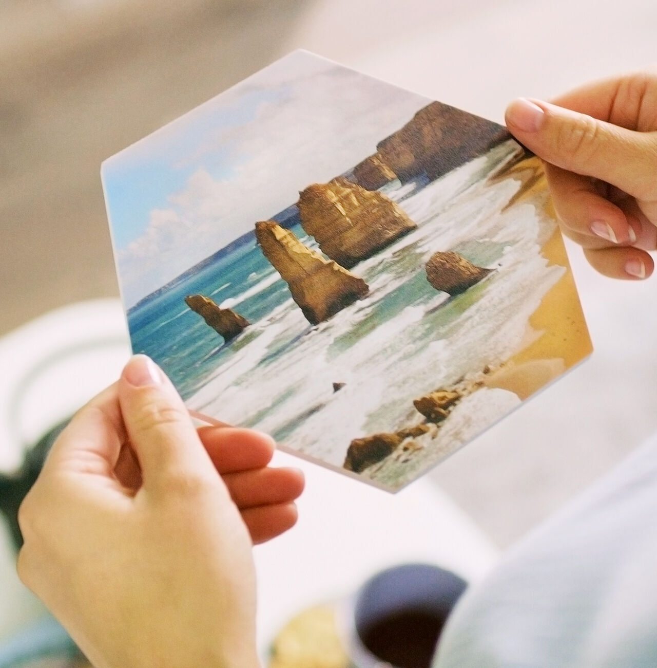 Iemand houdt een zeshoekige fotokaart vast met de beroemde Twelve Apostles aan de Australische kust. De oceaan en rotsen zijn goed zichtbaar.