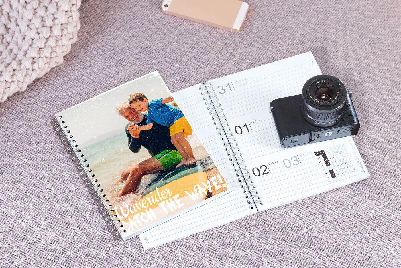 Een CEWE fotokalender ligt op een tafel naast een camera en een telefoon. Kalender toont een foto van een paar op het strand.