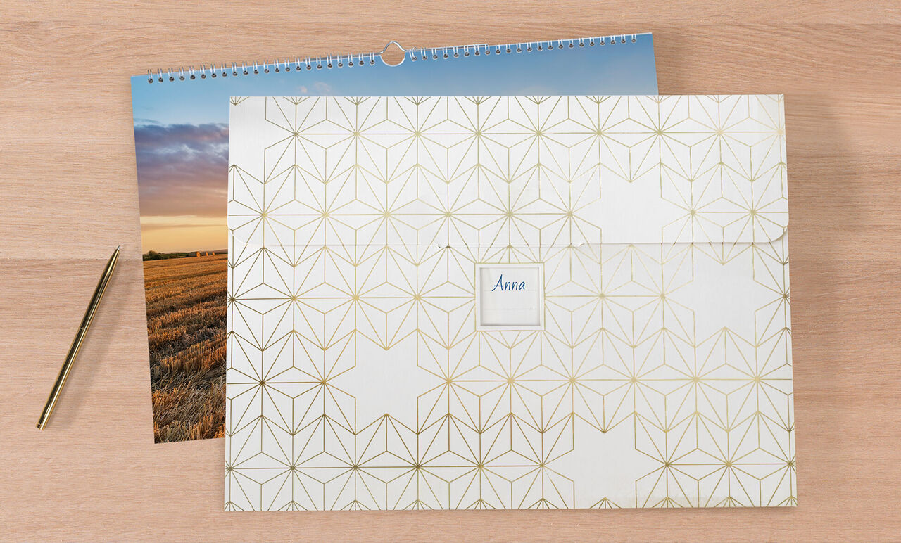 Een gesloten CEWE fotokalender op een houten oppervlak, naast een fotoafdruk en een gouden pen.