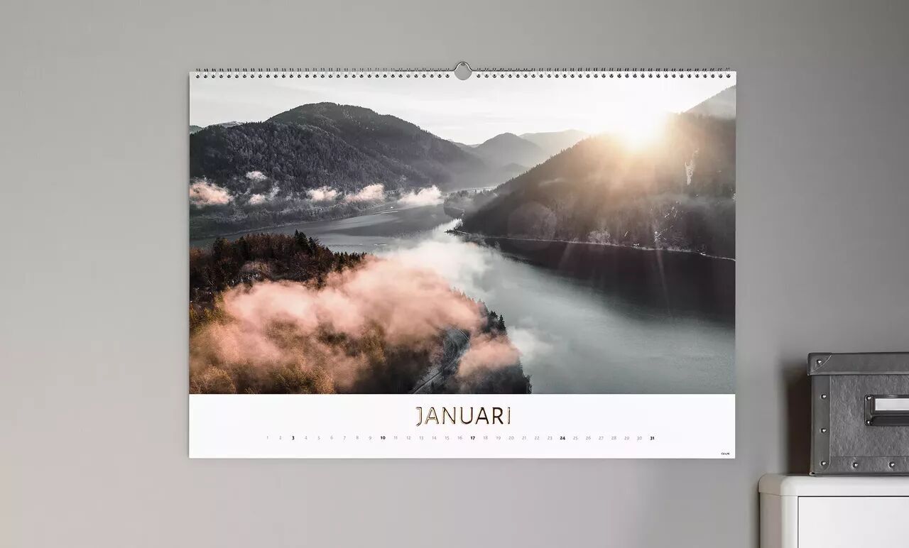 Een kalender hangt aan een muur. Hij toont een prachtig januarmotief met een schilderachtig landschap en zonnestralen.