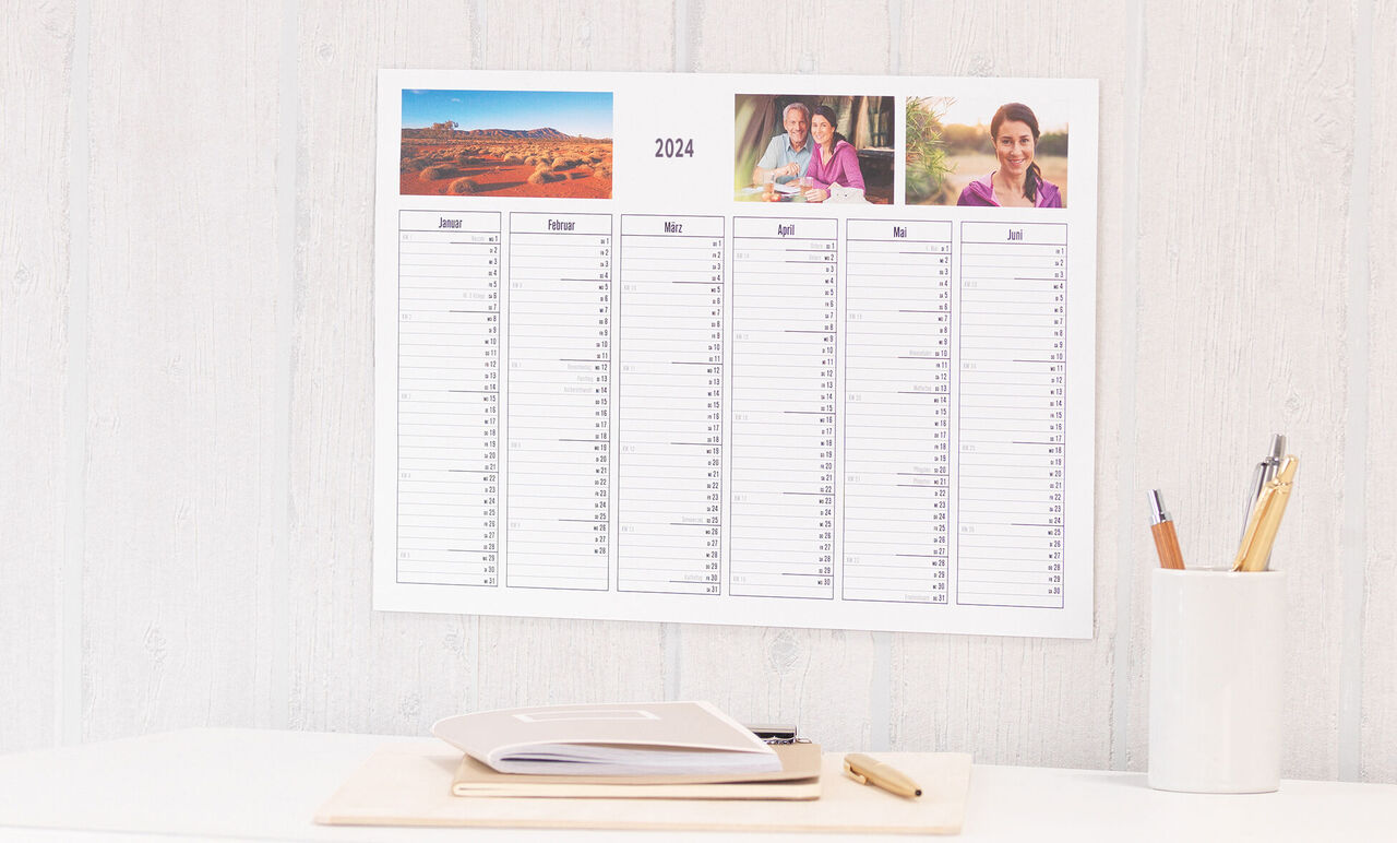 Een wandkalender voor 2024 hangt aan een witte muur. Ernaast liggen een notitieblok en pen op een tafel.