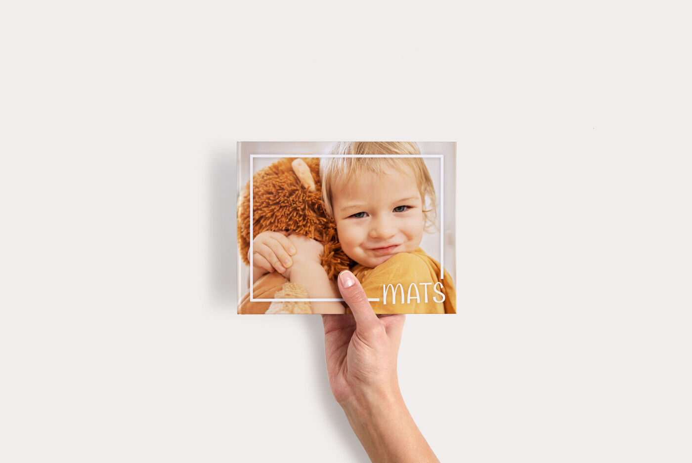 Klein fotoboek Compact Liggend met een kind dat een teddybeer knuffelt en de naam “Mats”, vastgehouden door één hand.