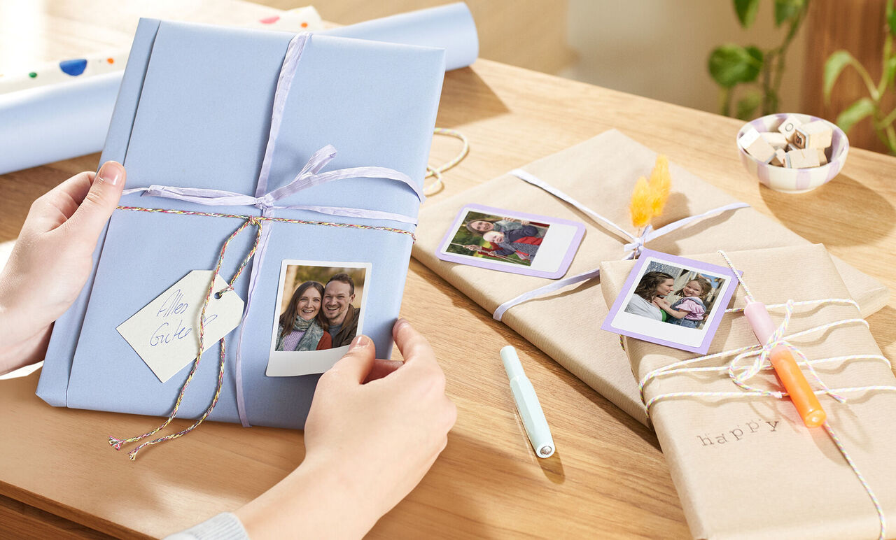 Une personne tient un cadeau emballé de manière créative avec des photos Polaroid dessus. D'autres cadeaux joliment emballés sont sur la table.