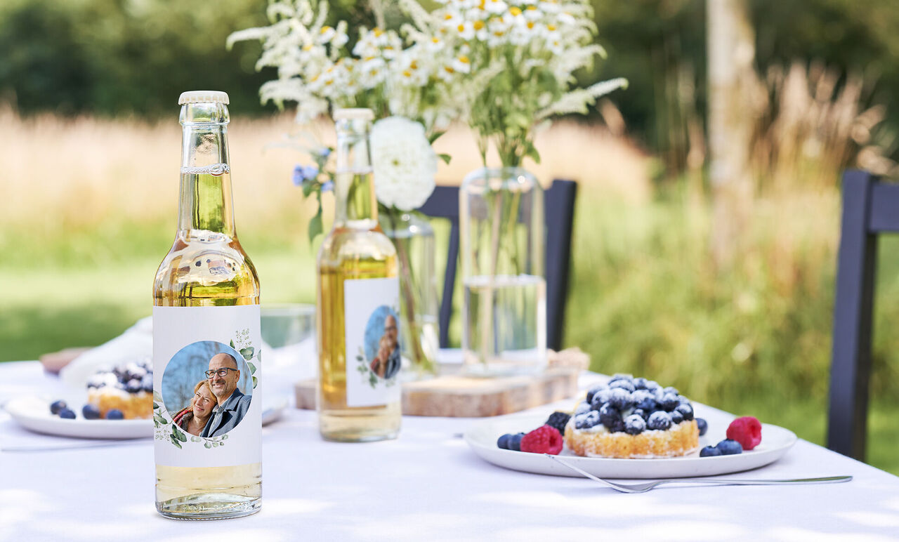 Deux bouteilles avec étiquettes personnalisées sur une table dressée, entourées de baies et d'un gâteau.