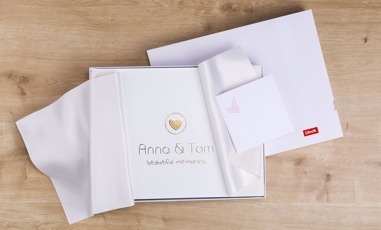 Un magnifique album photo CEWE avec une couverture beige lisse. Le couv. montre l'inscription "Anna & Tom" en lettres dorées avec un cœur.