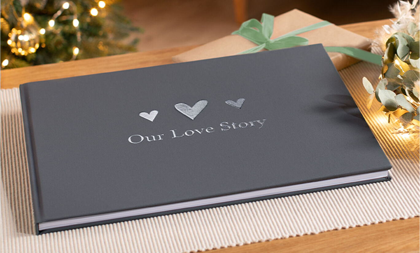Un beau CEWE ALBUM PHOTO intitulé 'Notre histoire d'amour'. Le livre est gris foncé et posé sur une table. En arrière-plan, on voit des cadeaux décorés avec soin et une ambiance festive.