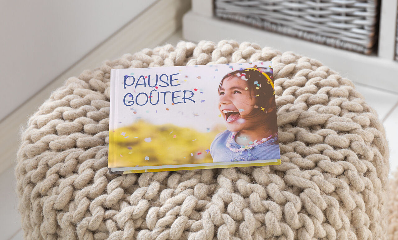 Un CEWE FOTOBUCH posé sur un pouf rond confortable avec une surface tricotée. La couverture dit "PAUSE DÉESSES" en lettres colorées.