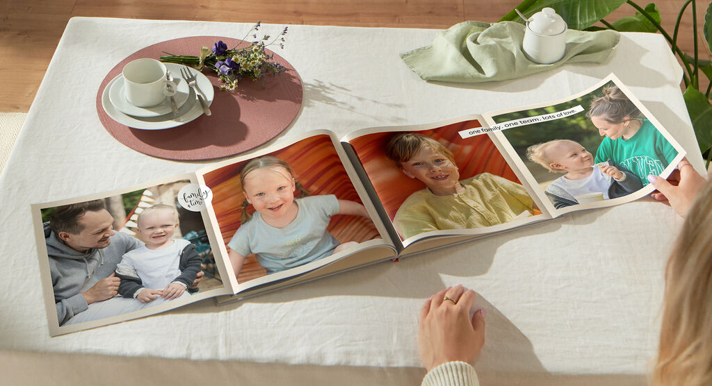 Une personne feuillette un album photo CEWE sur une table avec une tasse et un bouquet.