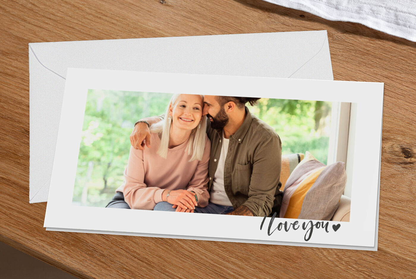 Un couple heureux assis sur un canapé, s'enlaçant avec le sourire. Une enveloppe marquée 'Love you' sur la table.